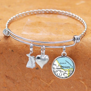 Puede incluir: Un brazalete de plata con tres abalorios: un corazón, un contorno del estado de Michigan y un mapa de Marquette, Michigan. El abalorio del mapa es azul, amarillo y verde.