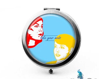 Puede incluir: Un espejo de bolsillo con fondo azul y dos caras estilizadas en rojo y amarillo. El texto "take your meds" está escrito en azul sobre las caras.