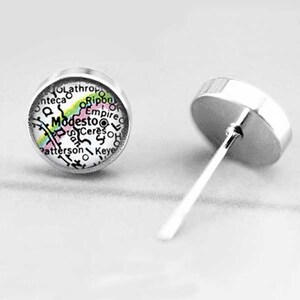 May include: A pair of silver stud earrings with a round, white, map-like design featuring the city of Modesto, California. The map includes the names of surrounding towns and cities.