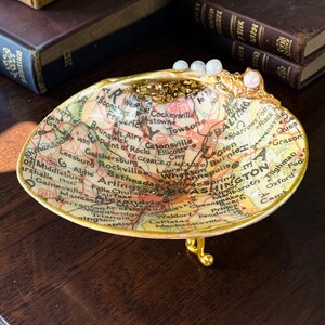 Puede incluir: Un cuenco decorativo con forma de concha que presenta un mapa antiguo de la zona de Washington D.C. y Baltimore. El interior está forrado con papel de mapa, acentuado con pan de oro y rematado con tres pequeñas perlas blancas sobre patas doradas.