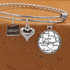 Puede incluir: Un brazalete de plata con tres dijes: un corazón, un mapa de Oregón y un mapa de Eugene, Oregón. Los dijes están unidos a un brazalete de plata.