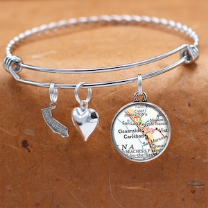 Puede incluir: Un brazalete de plata con tres dijes: un corazón, un contorno de California y un dije redondo con un mapa de Oceanside, California. El mapa muestra los nombres de las ciudades y pueblos cercanos, incluyendo Camp Pendleton, San Luis Rey de Francia, Vista y San Marcos.