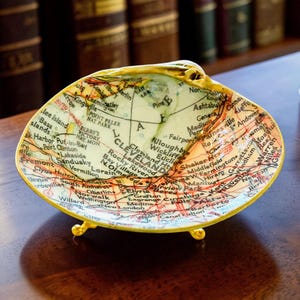Puede incluir: Un cuenco decorativo hecho de una concha marina grande, con un mapa en decoupage de la región de Cleveland, Ohio, en su superficie interior. La concha tiene un borde pintado de dorado y descansa sobre tres pequeñas patas de metal dorado.
