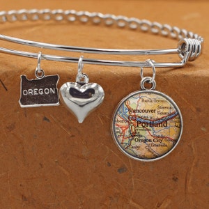 May include: A silver bangle bracelet with three charms: a heart, a map of Portland, Oregon, and a state outline of Oregon with the word "OREGON" inscribed.