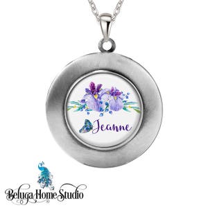 May include: A silver locket necklace with a white background and a watercolor design of purple irises and a blue butterfly. The locket has the name "Jeanne" written in purple lettering.