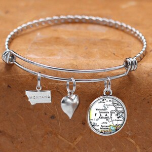 May include: A silver charm bracelet with three charms: a heart, a state outline of Montana, and a round charm with a map of Flathead Lake, Montana. The charms are attached to a silver chain bracelet.