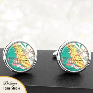 May include: A pair of silver cufflinks with a circular design featuring a vintage map of Oakland, California. The map is in shades of green, yellow, and red, with the city name prominently displayed.