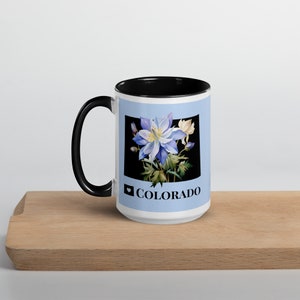 Könnte beinhalten: Weiße Keramiktasse mit schwarzem Rand und Henkel. Die Tasse zeigt eine blaue und weiße Blumenillustration mit dem Text "♥ COLORADO" auf der Vorderseite.