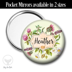 May include: A round pocket mirror with a floral design featuring a variety of flowers, including clover, daisies, and lavender. The mirror has a white background with a peach-colored frame and the name "Heather" written in black script.