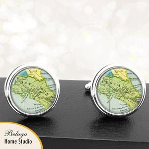 May include: A pair of silver cufflinks with a round, glass-like surface featuring a vintage map of Costa Rica. The map is in shades of green, yellow, and brown, with the text "COSTA RICA" printed in black.