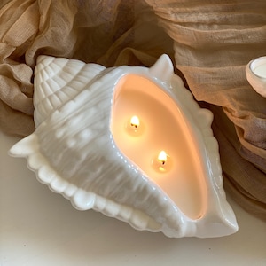 Puede incluir: Un portavelas blanco en forma de concha con dos velas encendidas. El portavelas tiene un acabado brillante y está relleno de cera cremosa. Las llamas proyectan un brillo cálido. El fondo presenta una tela beige.