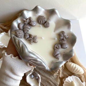 May include: A clam shell-shaped candle holder with a silver trim, filled with a creamy white candle. Decorative silver seashells and starfish decorate the candle's surface. A unique home decor item.