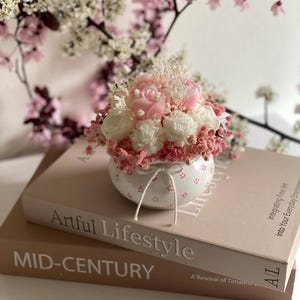 Può includere: Un piccolo vaso di ceramica bianca con una composizione floreale rosa, tra cui peonie e altri fiori, è posto sopra due libri. I libri sono intitolati "Artful Lifestyle" e "MID-CENTURY". Lo sfondo presenta fiori rosa e bianchi.