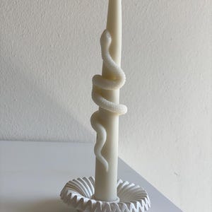 Peut inclure: Une longue bougie effilée blanc cassé avec un serpent sculpté enroulé autour de son corps. La bougie est maintenue droite dans un bougeoir circulaire blanc doté d'un bord décoratif plissé ou volanté, créant un aspect texturé.