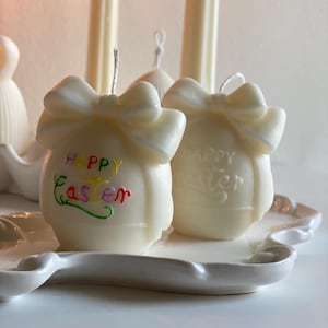May include: Two ivory-colored Easter egg-shaped candles with bow accents. One candle has the words "HAPPY Easter" in colorful script. The candles sit on a white, scalloped-edge tray, with additional candles in the background.