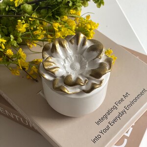 May include: A white ceramic trinket box with a flower-shaped lid, accented with gold trim. The box is sitting on a stack of books with the text "Integrating Fine Art into Your Everyday Environment". Yellow and green floral arrangement in the background.
