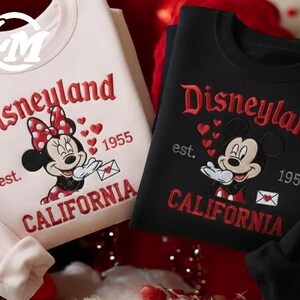 two sweatshirts with Disney characters and text, one in white and one in black, both with the text "Disneyland est. 1955" and "California" printed on them.
