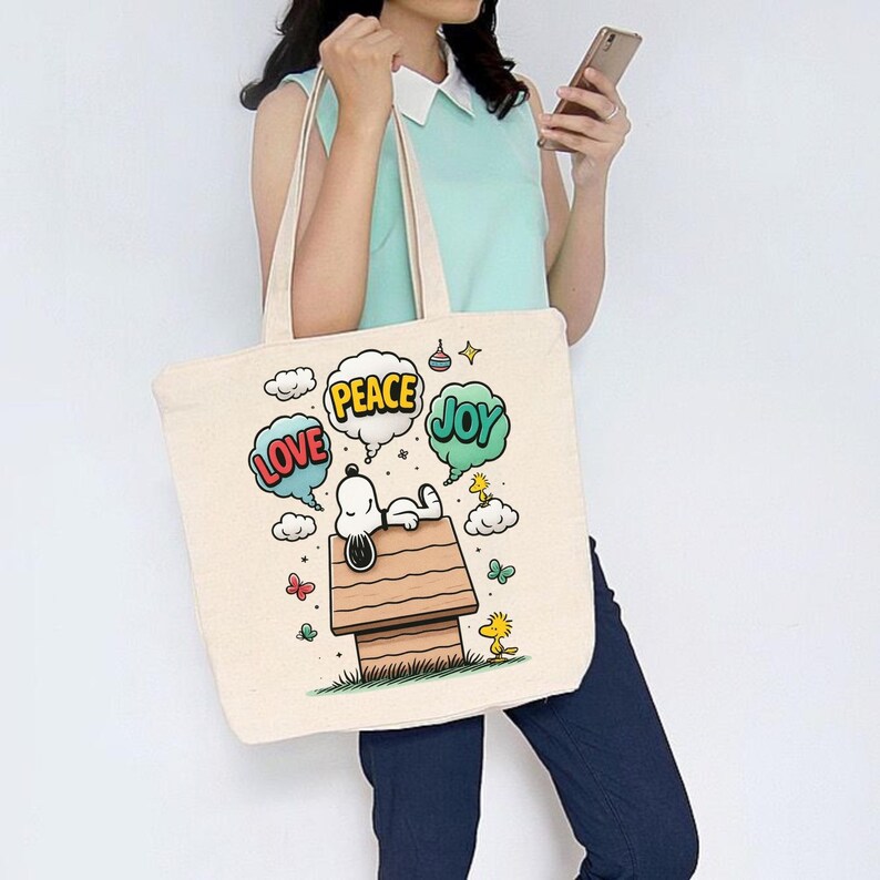 May include: Cream-colored tote bag featuring a Snoopy graphic. Snoopy is depicted relaxing on his doghouse, with speech bubbles that say "Love", "Peace", and "Joy". The bag has two beige handles.