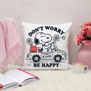 May include: White square pillow featuring Snoopy driving a car, holding a flower, with the text "DON'T WORRY BE HAPPY". The pillow also has a flower and a small red planter on the car. A cute and cheerful design.