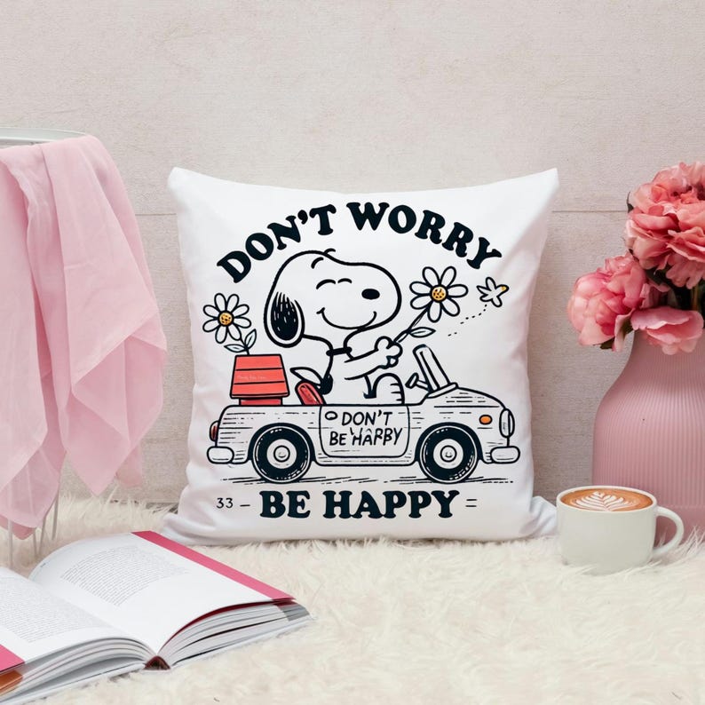 May include: White square pillow featuring Snoopy driving a car, holding a flower, with the text "DON'T WORRY BE HAPPY". The pillow also has a flower and a small red planter on the car. A cute and cheerful design.