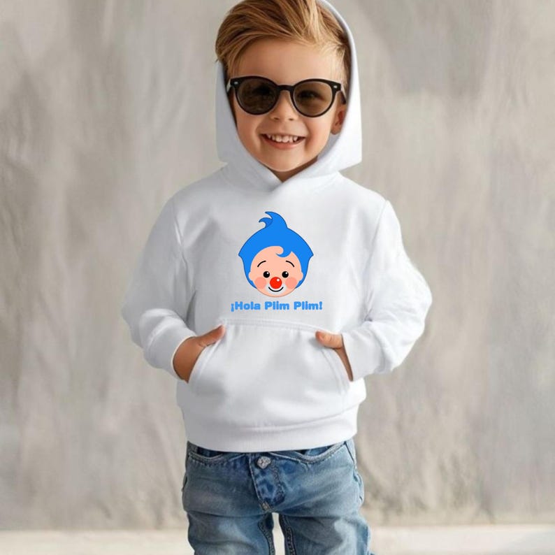 May include: White hooded sweatshirt with a cartoon character design, blue hair, a red nose, and the text "&iexcl;Hola Plim Plim!" in blue. The sweatshirt has a front pocket and is worn by a child wearing sunglasses and blue jeans.