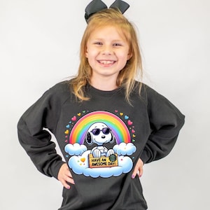 May include: A black sweatshirt featuring a colorful rainbow and a cartoon dog wearing sunglasses. The dog is sitting on a cloud and holding a sign that says "Have an Awesome Day!" The design is surrounded by hearts.