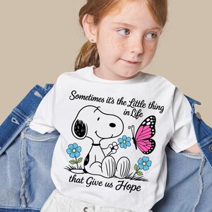 May include: White t-shirt featuring a cartoon Snoopy character holding a flower, with a pink butterfly and blue flowers. The shirt has the text "Sometimes it's the Little thing in Life that Give us Hope".