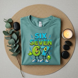 May include: A teal t-shirt with the words "SIX SEVEN" in a gradient blue and green font, with lightning bolt accents. Below, cartoon numbers 6 and 7 with faces and sneakers. The shirt is on a woven mat with a candle and decorative stones.