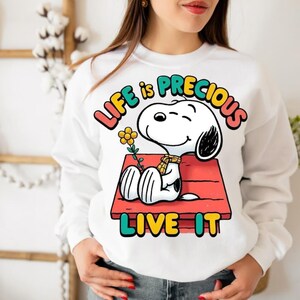 May include: White sweatshirt featuring a colorful graphic of Snoopy from Peanuts, resting on his doghouse with a flower. The text "LIFE is PRECIOUS LIVE IT" is displayed in vibrant colors. A casual and comfortable apparel item.