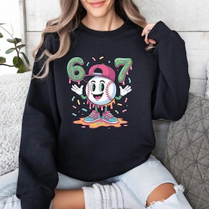 May include: Black sweatshirt featuring a baseball character with a smiling face, wearing a pink cap and sneakers. The number "67" in green frosting is above the baseball character. Colorful sprinkles are scattered around the design.