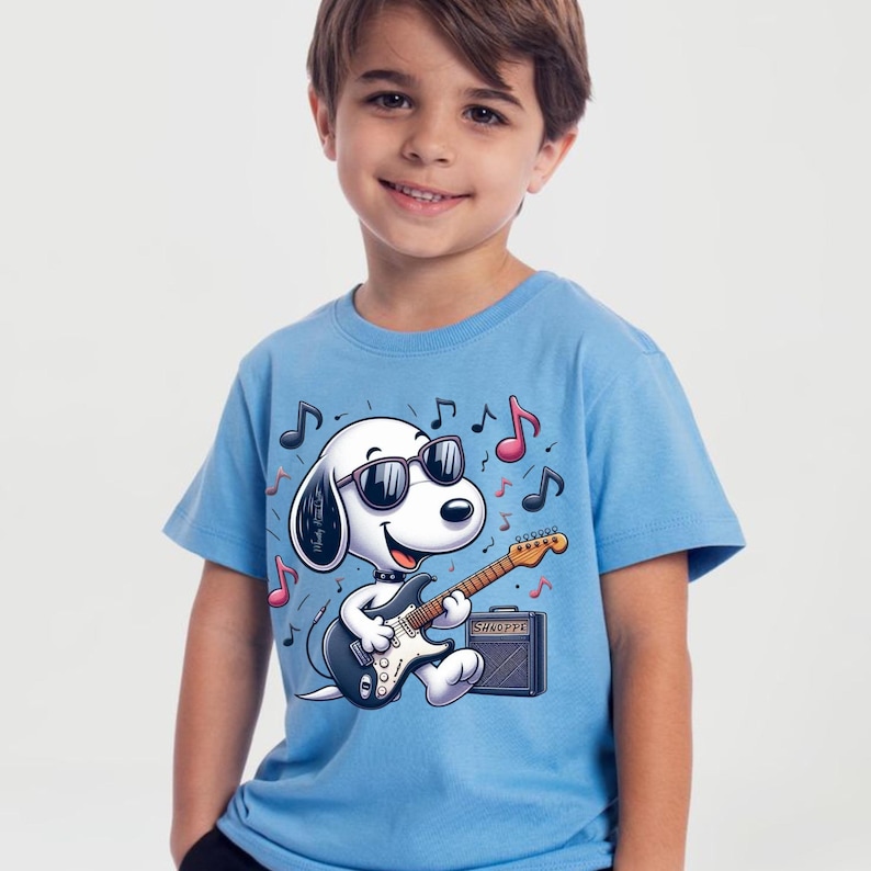 May include: Light blue t-shirt featuring a cartoon dog wearing sunglasses and playing a black electric guitar. The dog is in front of a gray amplifier, with musical notes scattered around. The word "SHOPPE" is on the amplifier.