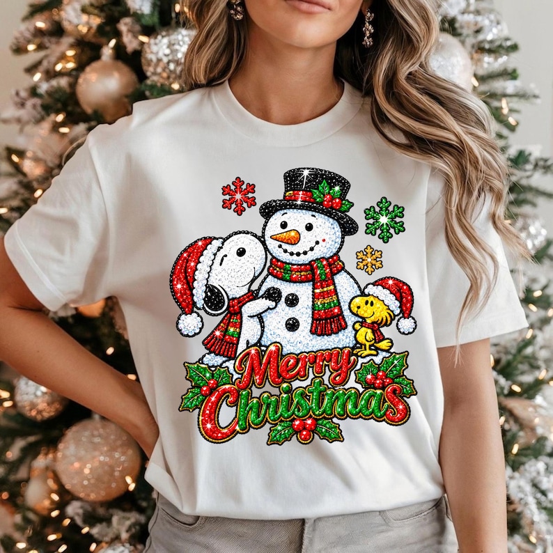 May include: A white t-shirt featuring a graphic print of Snoopy and Woodstock wearing Santa hats next to a snowman with a scarf and top hat. The design includes red and green snowflakes, holly berries, and the text Merry Christmas in red and green.