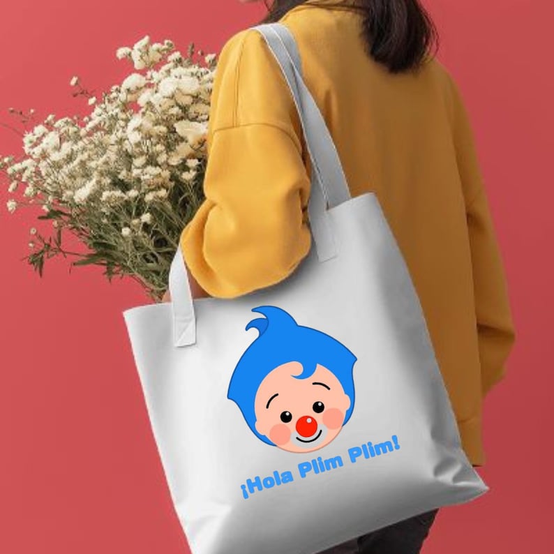 May include: White tote bag with a cartoon face with blue hair, a red nose, and the text "&iexcl;Hola Plim Plim!" in blue. The bag has white handles and is held by a person wearing a yellow sweatshirt. A bouquet of white flowers is also visible.