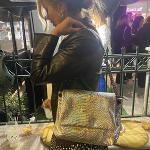 Peut inclure: Un sac à bandoulière rectangulaire doté d'une finition métallique irisée à texture peau de serpent. Le sac présente un rabat structuré et une chaîne sombre. Le matériau reflète la lumière avec des teintes arc-en-ciel changeantes.