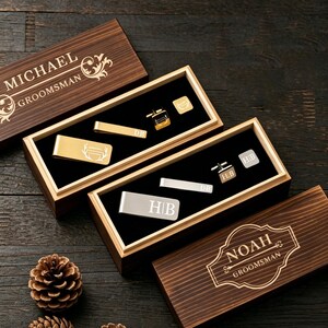 May include: Wooden gift boxes with personalized text, containing gold and silver cufflinks, tie clips, and money clips. The boxes are engraved with names and the word "Groomsman". The items are displayed on a black velvet surface.