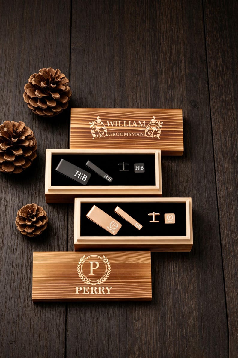 May include: Wooden gift boxes with engraved text and accessories. The boxes contain tie clips, cufflinks, and money clips. The text on the boxes reads "WILLIAM GROOMSMAN" and "PERRY". The accessories are black and gold.