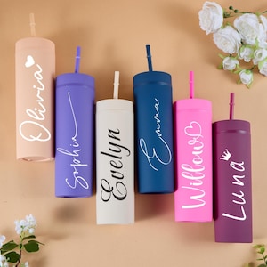 May include: Six cylindrical tumblers with straws in peach, purple, cream, navy blue, pink, and dark plum. Each tumbler features a personalized name in white or black script: Olivia, Sophia, Evelyn, Emma, Willow, and Luna, with small icons.
