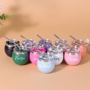 Peut inclure: Une collection de dix gobelets ronds en acier inoxydable de différentes couleurs, dont vert, rose, blanc, noir, violet et orange. Chaque gobelet possède un couvercle transparent, une paille métallique courbée et un prénom personnalisé en blanc.