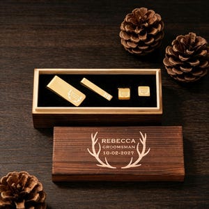 May include: A wooden box set with gold-colored accessories, including a tie clip, cufflinks, and a money clip. The box is engraved with the name "Rebecca", the word "Groomsman", and the date "10-02-2027". Pine cones are scattered around the box.