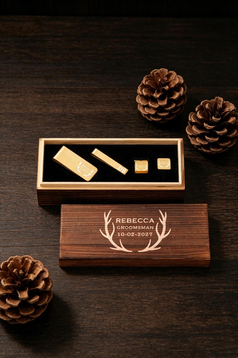 May include: A wooden box set with gold-colored accessories, including a tie clip, cufflinks, and a money clip. The box is engraved with the name "Rebecca", the word "Groomsman", and the date "10-02-2027". Pine cones are scattered around the box.