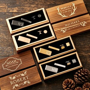 May include: Wooden gift boxes with personalized groomsmen gifts, including money clips, tie clips, and cufflinks. The boxes are engraved with names and dates. The gifts are displayed on a black velvet lining. The overall color scheme is brown, gold, and silver.