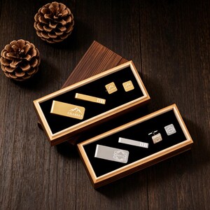 May include: Two wooden boxes with black velvet interiors display gold and silver accessories. The gold set includes a money clip, tie bar, and cufflinks. The silver set features a money clip, tie bar, and cufflinks. Pinecones rest on a dark wood surface.