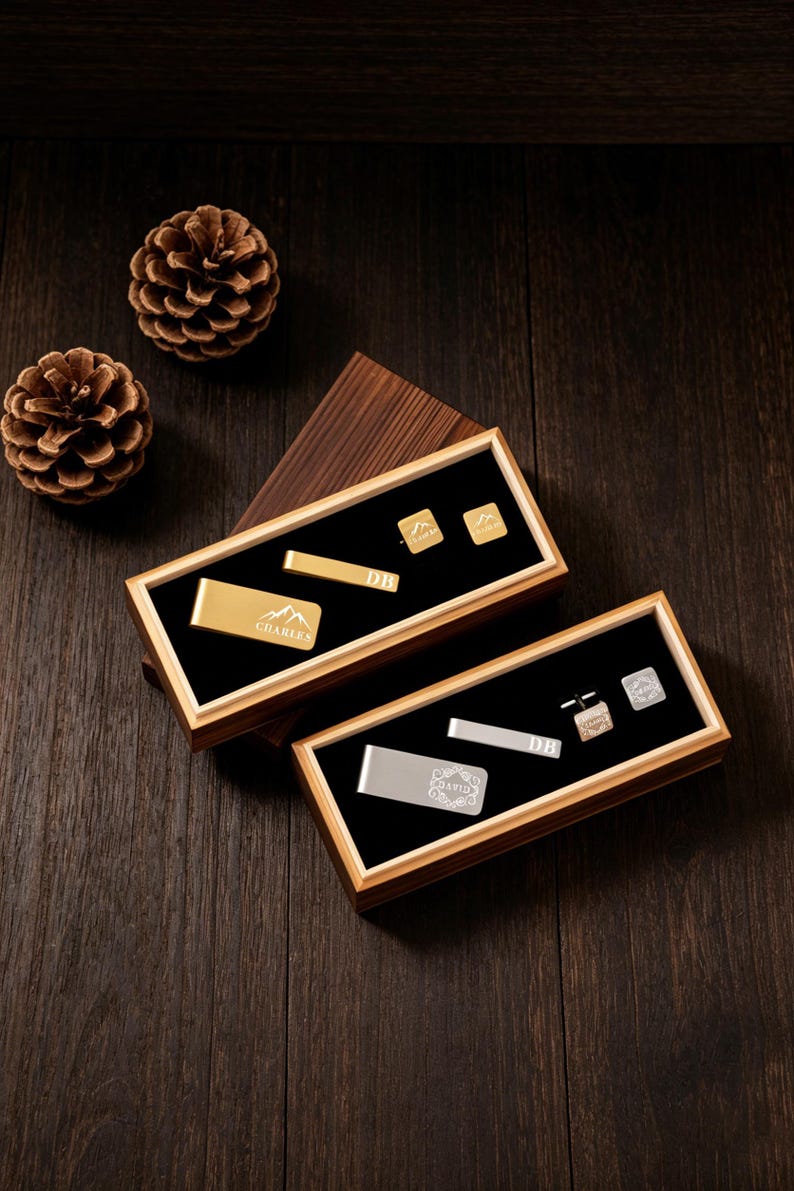 May include: Two wooden boxes with black velvet interiors display gold and silver accessories. The gold set includes a money clip, tie bar, and cufflinks. The silver set features a money clip, tie bar, and cufflinks. Pinecones rest on a dark wood surface.