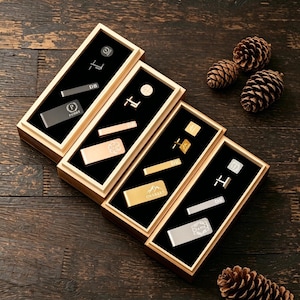 May include: An overhead shot of four wooden boxes, each containing various men's accessories. The accessories include tie clips, cufflinks, and money clips in silver and gold tones, with personalized initials and designs. The boxes are on a dark wood surface.