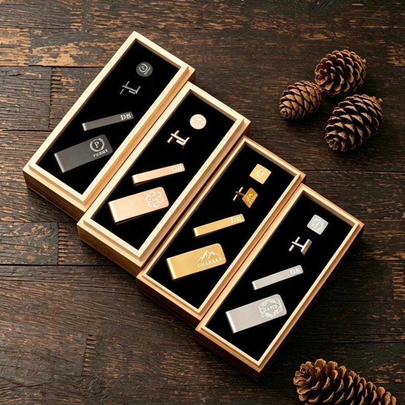 May include: An overhead shot of four wooden boxes, each containing various men's accessories. The accessories include tie clips, cufflinks, and money clips in silver and gold tones, with personalized initials and designs. The boxes are on a dark wood surface.