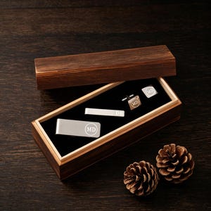 May include: A wooden box set containing a silver money clip, tie bar, and cufflinks. Each piece is engraved with the initials "MD" in a circle. The tie bar is engraved with "DB". The box is open, revealing the items on a black velvet lining.
