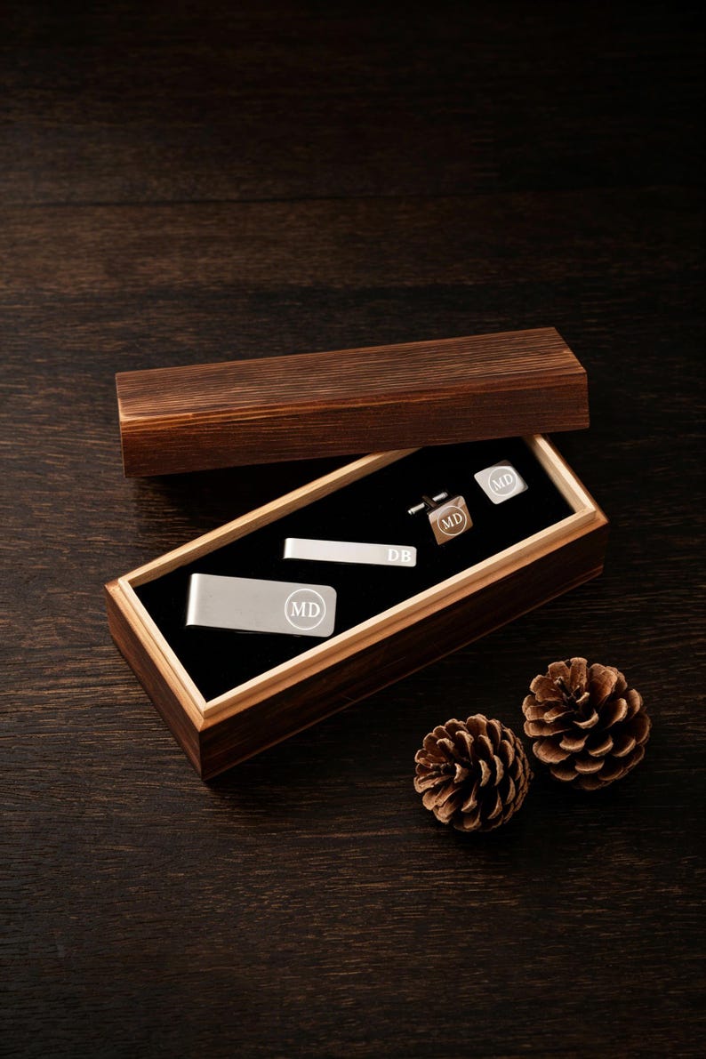 May include: A wooden box set containing a silver money clip, tie bar, and cufflinks. Each piece is engraved with the initials "MD" in a circle. The tie bar is engraved with "DB". The box is open, revealing the items on a black velvet lining.