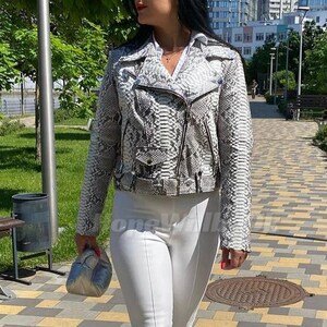 May include: A woman wearing a snakeskin-patterned jacket, white pants, and platform sneakers walks on a paved path. The jacket is gray and white with a zipper closure. She carries a small silver purse. The path is made of yellow, gray, and brown tiles.