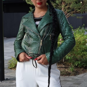Può includere: Giacca in pelle verde con motivo a pelle di serpente, con cintura in vita e cerniere dorate. La giacca ha un classico stile biker con colletto a revers e maniche lunghe. La modella indossa pantaloni bianchi e un top bianco e nero.