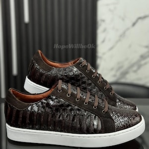 May include: A pair of brown leather sneakers with a crocodile skin pattern. The shoes have brown suede accents, brown laces, and white soles. The sneakers are displayed on a reflective surface.
